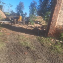 MagBud Rozbiórki,Wyburzenia i Prace ziemne Magdalena Musiał - Żółta koparko-ładowarka na placu budowy, fragment ceglanej konstrukcji w tle, ślady opon na ziemi, błękitne niebo.