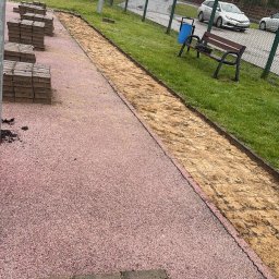 MagBud Rozbiórki,Wyburzenia i Prace ziemne Magdalena Musiał - Przygotowanie terenu pod chodnik z kostki brukowej na zewnątrz. Ułożone kostki brukowe czekają na montaż. Widoczny fragment trawnika i ogrodzenia.