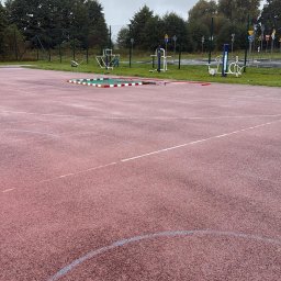 MagBud Rozbiórki,Wyburzenia i Prace ziemne Magdalena Musiał - Czerwona nawierzchnia boiska z liniami, w tle urządzenia do ćwiczeń na świeżym powietrzu, ogrodzenie i drzewa. Pochmurny dzień.
