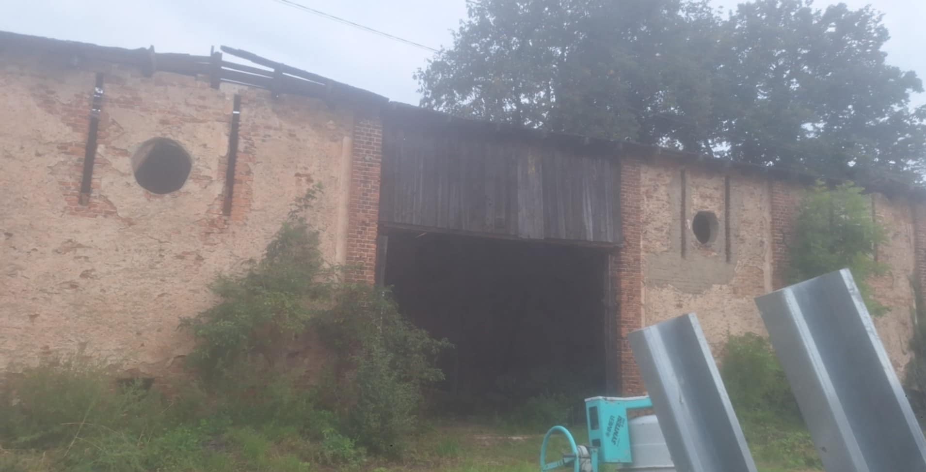 Stary, ceglano-kamienny budynek z drewnianą bramą i okrągłymi otworami w ścianie, częściowo zrujnowany, z widoczną zielenią i sprzętem budowlanym.