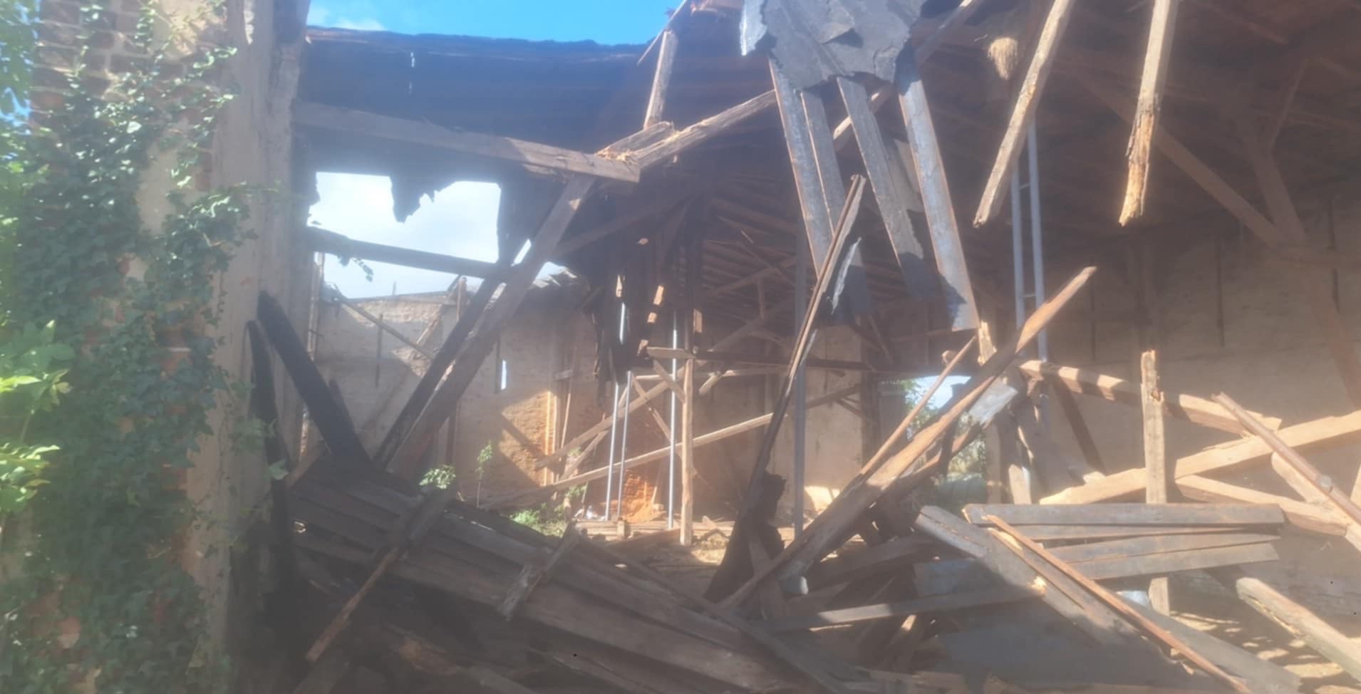 Zdjęcie przedstawia zniszczony budynek w trakcie rozbiórki. Widoczne elementy konstrukcyjne, uszkodzony dach i ściany. Fragmenty roślinności na murze. Niebo w tle.