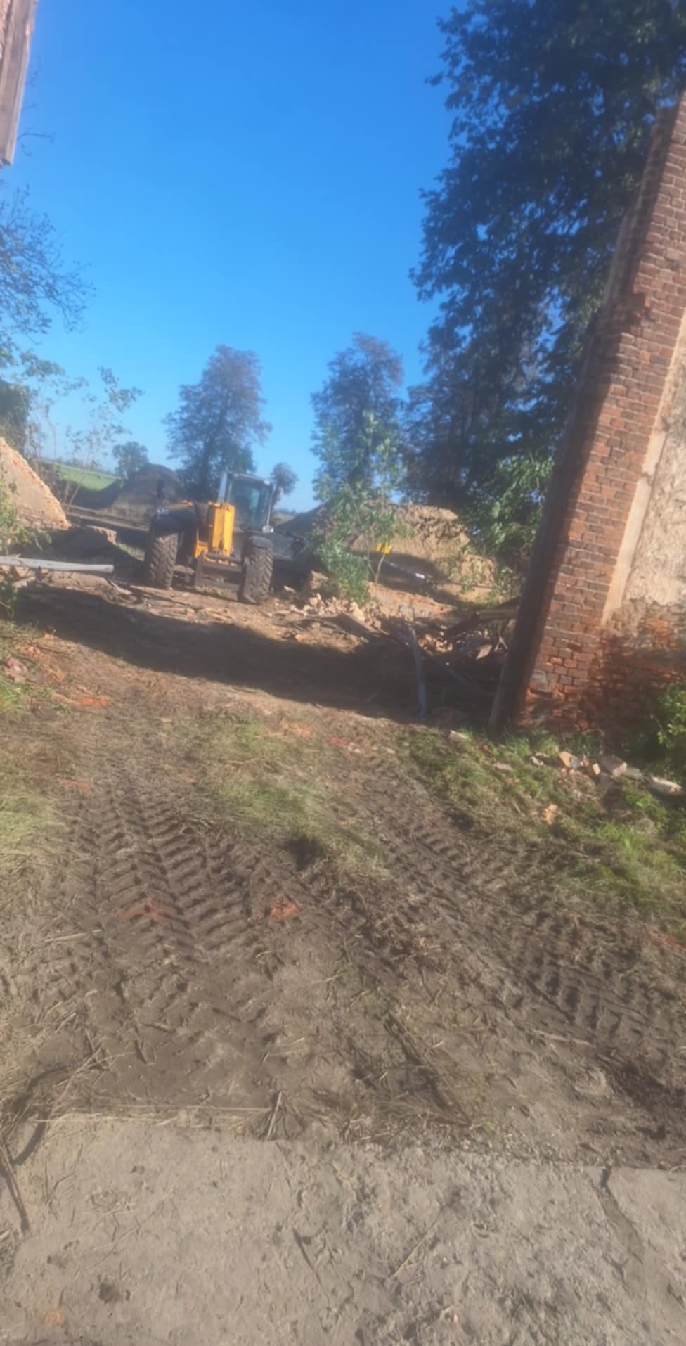 Żółta koparko-ładowarka na placu budowy, fragment ceglanej konstrukcji w tle, ślady opon na ziemi, błękitne niebo.