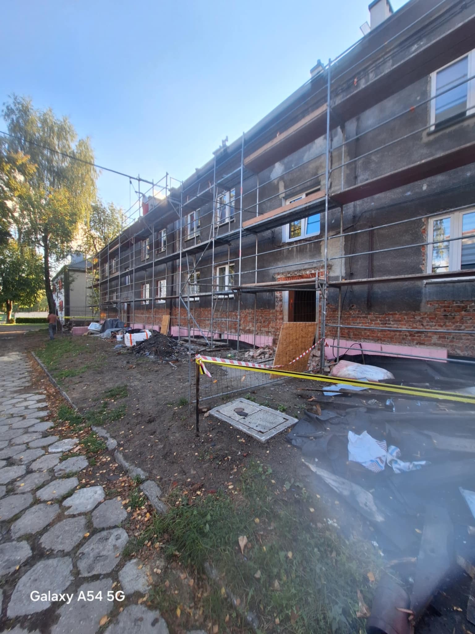 Budynek w trakcie remontu z rusztowaniami, widoczne ocieplenie, częściowo odsłonięta cegła. Na pierwszym planie gruz i taśma ostrzegawcza.