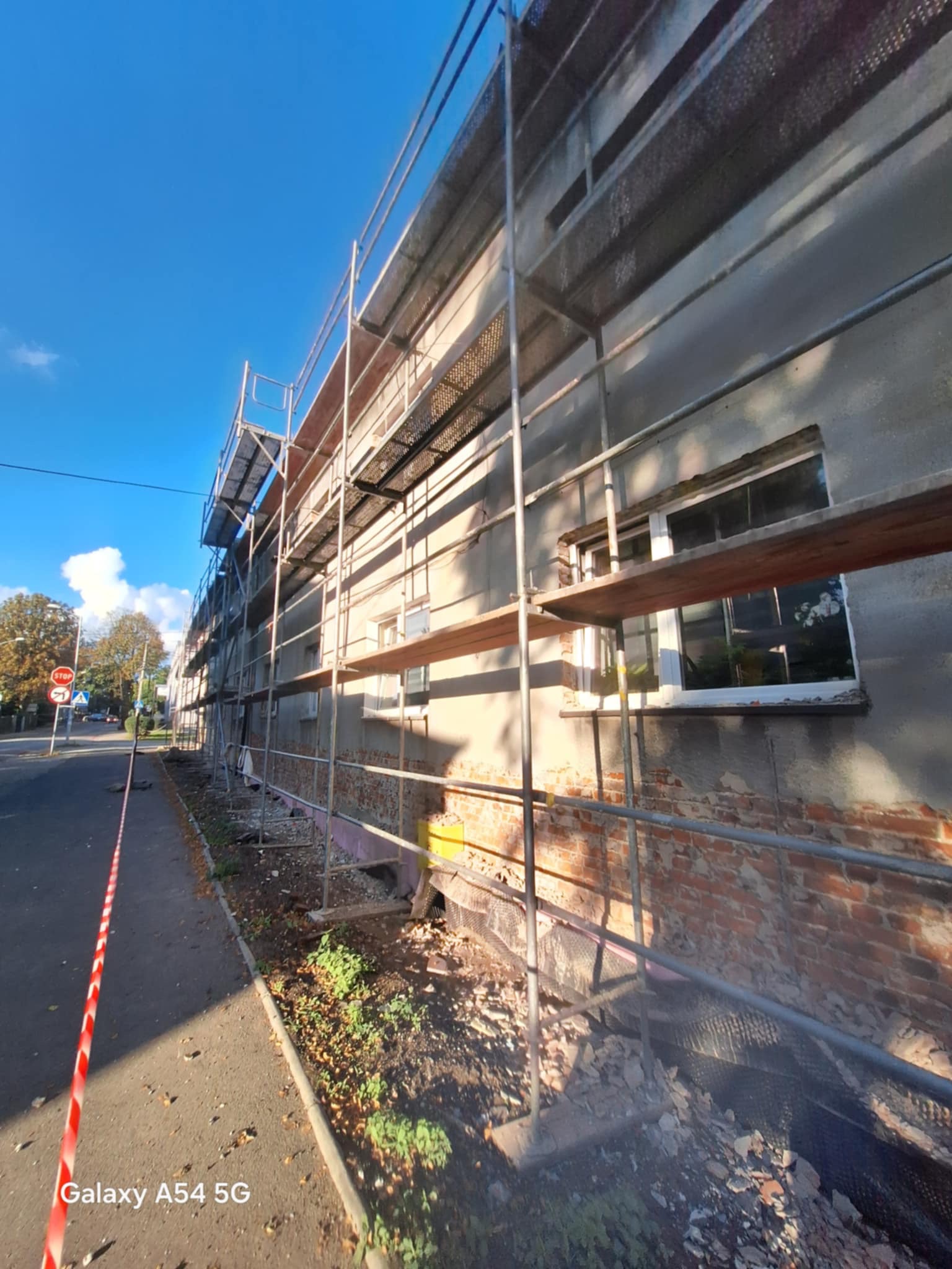 Budynek w trakcie remontu z rusztowaniem. Częściowo odsłonięta cegła, widoczne okna i niebieskie niebo. Ujęcie z dołu, z perspektywy ulicy.