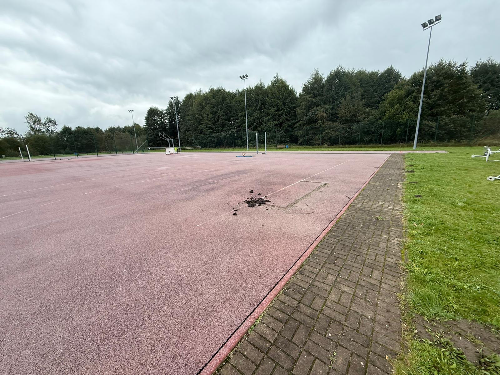 Czerwona nawierzchnia boiska sportowego z widocznymi śladami naprawy, otoczona zielenią i oświetlona przez wysokie lampy, pod pochmurnym niebem.