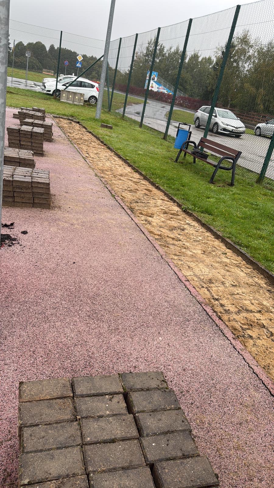 Przygotowanie terenu pod chodnik z kostki brukowej na zewnątrz. Ułożone kostki brukowe czekają na montaż. Widoczny fragment trawnika i ogrodzenia.