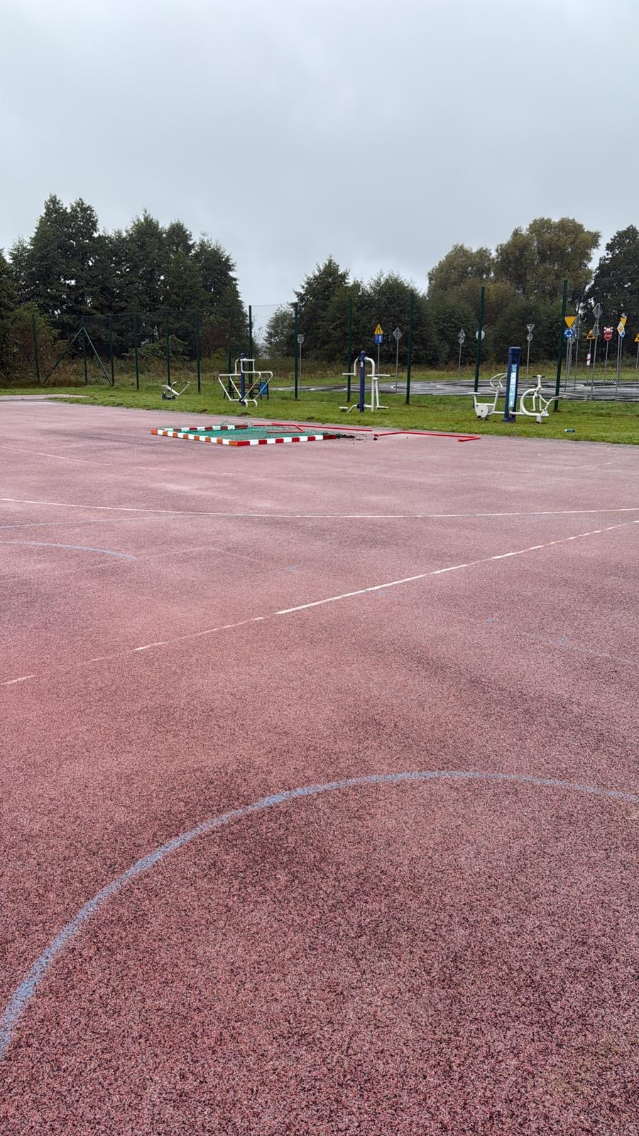Czerwona nawierzchnia boiska z liniami, w tle urządzenia do ćwiczeń na świeżym powietrzu, ogrodzenie i drzewa. Pochmurny dzień.