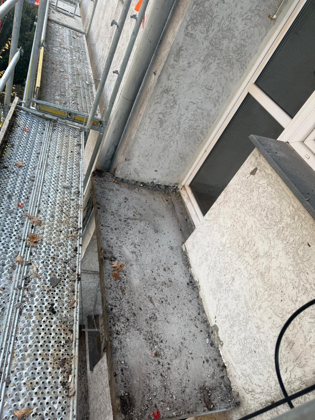 Balkon w trakcie remontu z rusztowaniem. Widoczne ślady prac, okno i fragment elewacji z fakturą. Ujęcie z góry, podkreślające etap prac wykończeniowych.