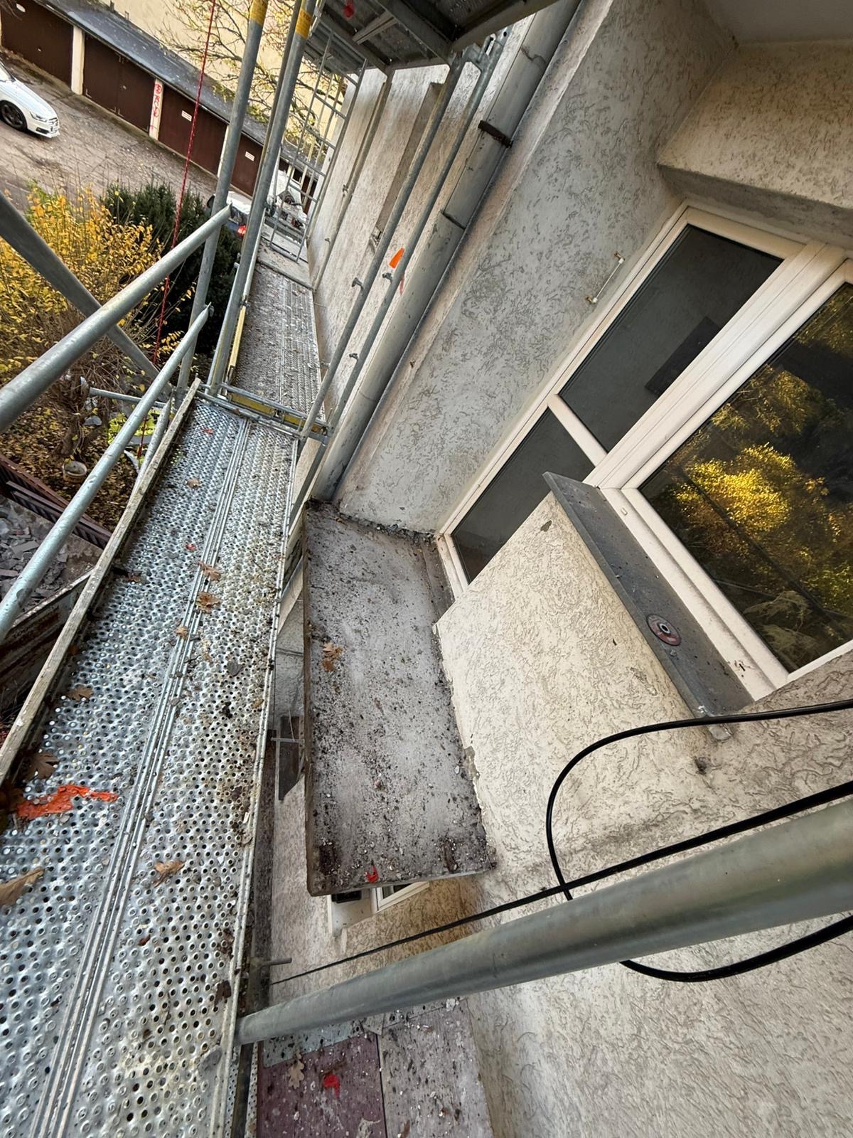 Elewacja budynku w trakcie prac remontowych, widok z rusztowania. Okno z białymi ramami, szara ściana z fakturą. Elementy rusztowania w kadrze.