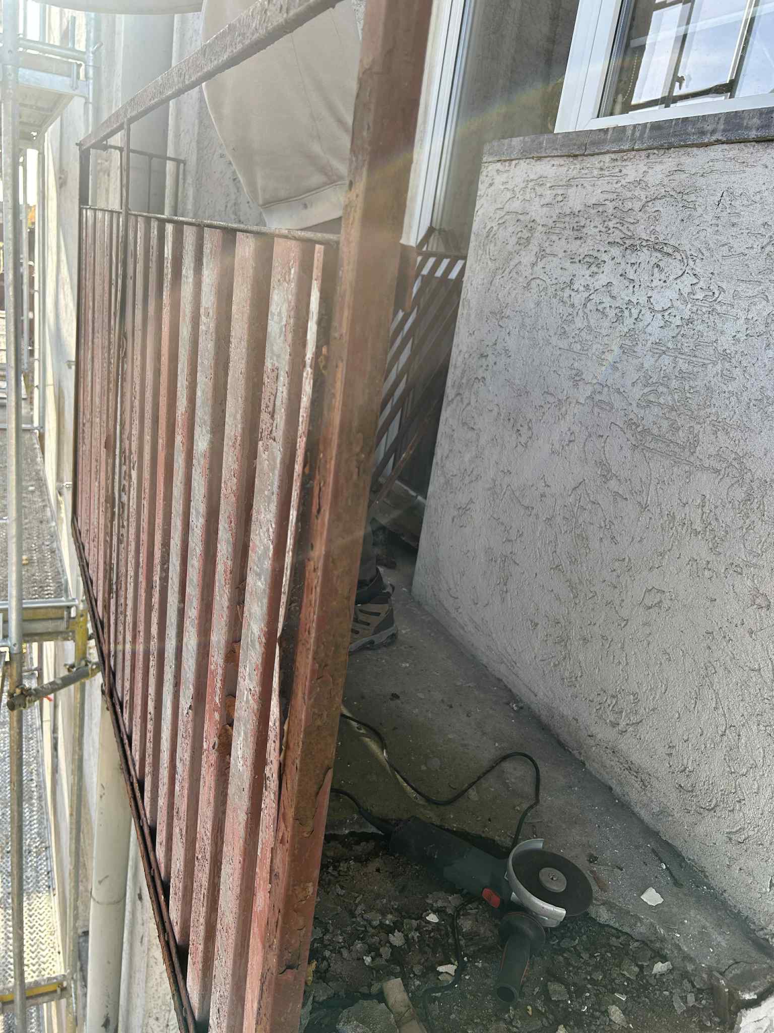 Demontaż starej, zardzewiałej balustrady balkonowej z użyciem szlifierki kątowej. Widoczne rusztowanie i fragment elewacji z fakturą. Prace rozbiórkowe.