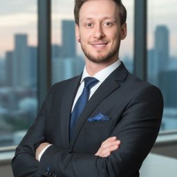 Północ Nieruchomości - Uśmiechnięty mężczyzna w garniturze z niebieskim krawatem i poszetką, stojący na tle okna z widokiem na miasto. Skrzyżowane ręce, kolczyk w uchu.