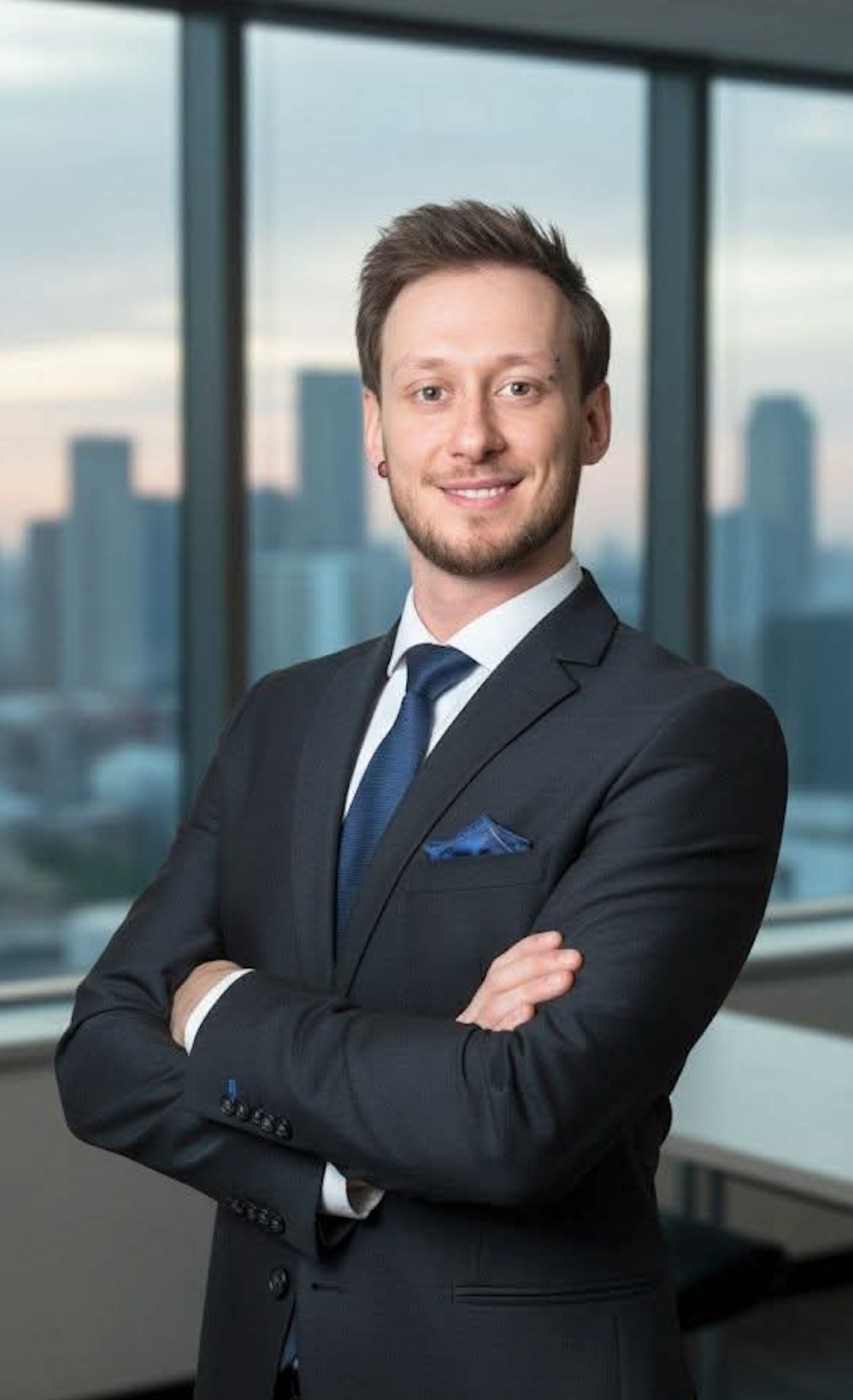 Uśmiechnięty mężczyzna w garniturze z niebieskim krawatem i poszetką, stojący na tle okna z widokiem na miasto. Skrzyżowane ręce, kolczyk w uchu.