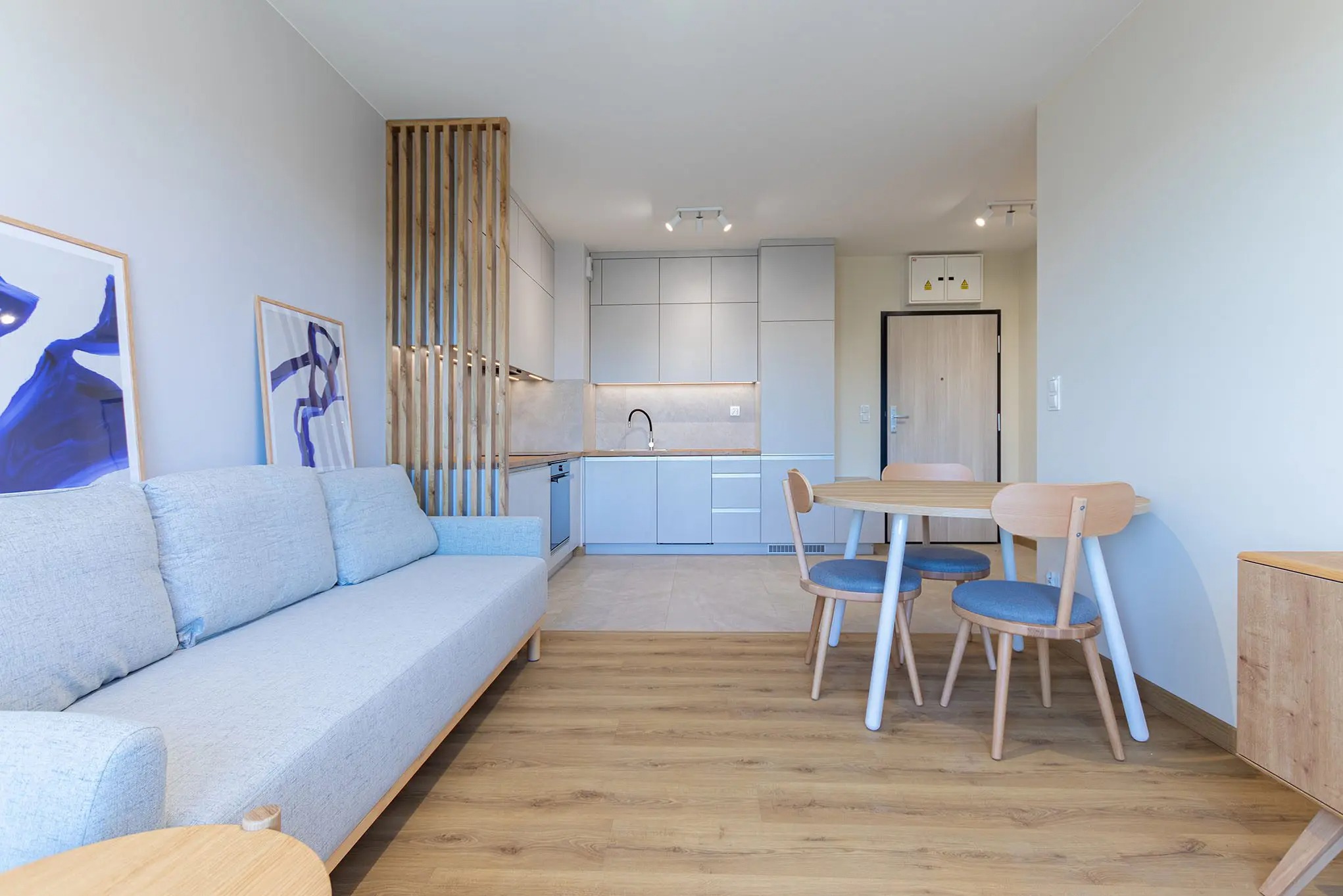 Jasne, nowoczesne wnętrze po remoncie w Krakowie: salon z sofą, jadalnia i aneks kuchenny oddzielony drewnianą ścianką. Parkiet na podłodze.