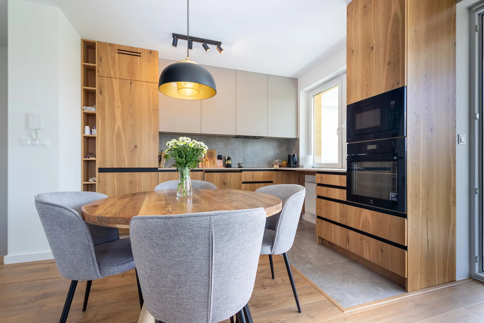 Nowoczesna kuchnia z drewnianymi szafkami, okrągłym stołem i szarymi krzesłami. Czarna lampa wisząca nad stołem. Widok na okno i blat kuchenny.
