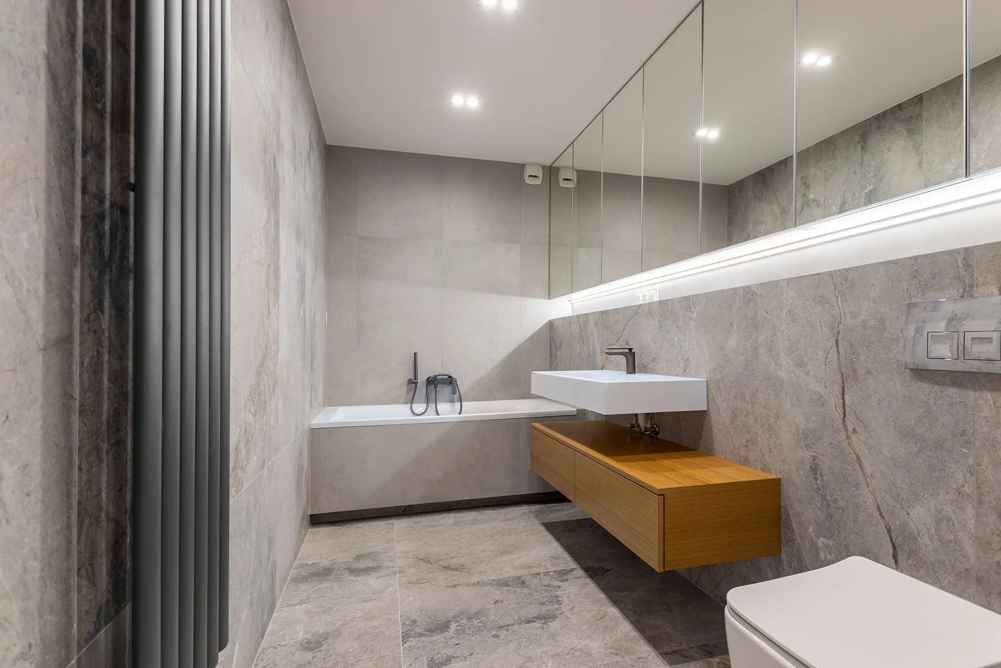 Nowoczesna łazienka z płytkami imitującymi marmur, podwieszaną szafką z umywalką i grzejnikiem żeberkowym. Minimalistyczny design z lustrami na ścianie.