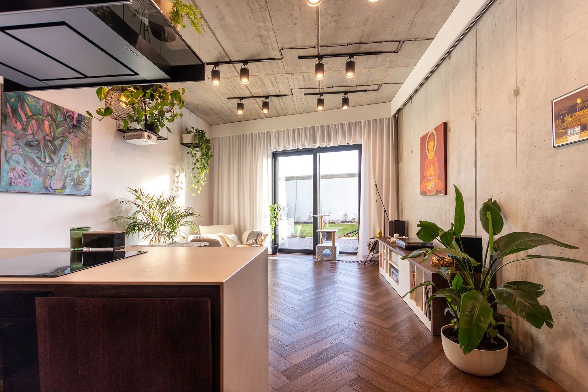 Nowoczesne wnętrze z parkietem w jodełkę, surowym betonem na ścianach i suficie oraz licznymi roślinami doniczkowymi. Widoczna część kuchni i wyjście na taras.