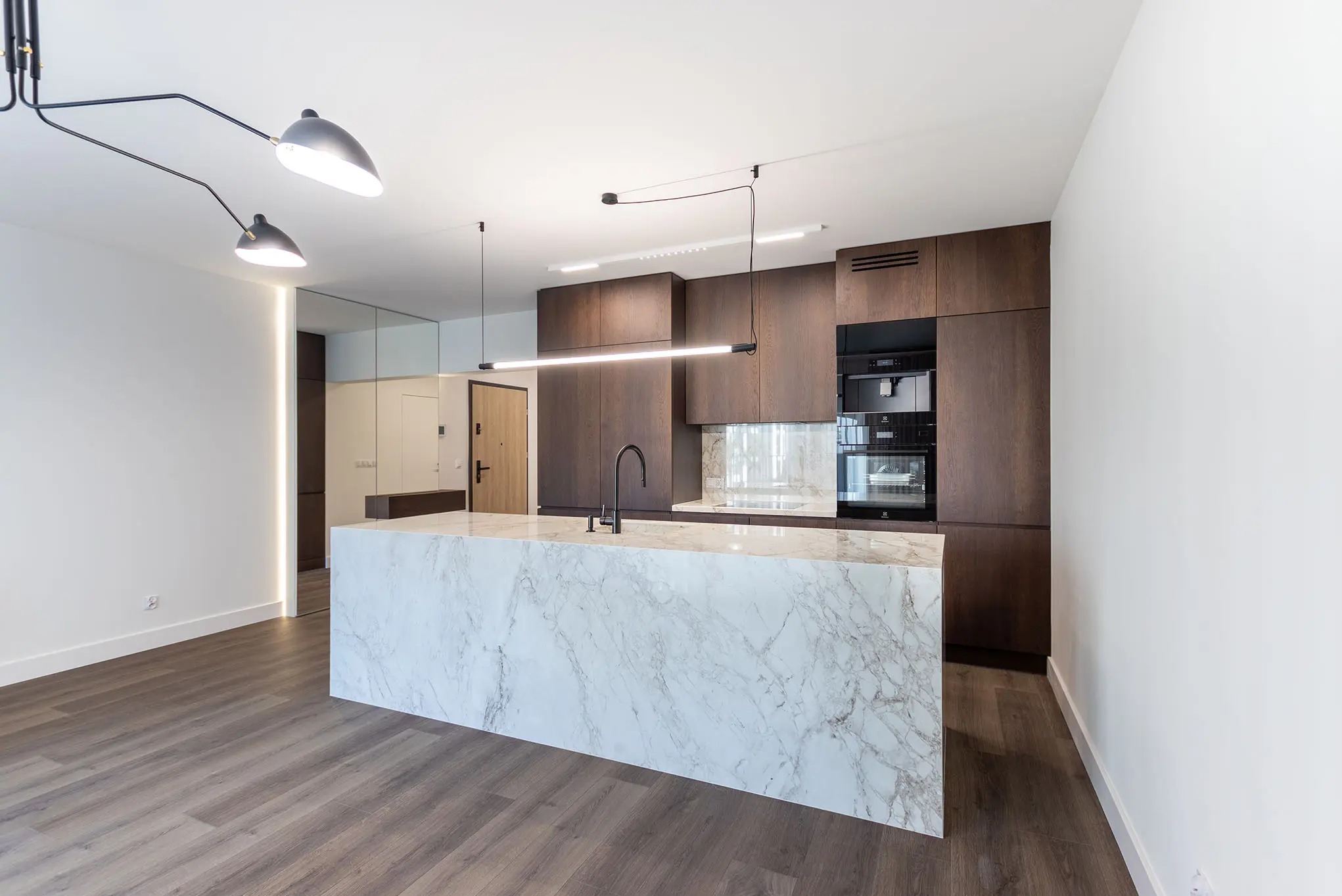Nowoczesna kuchnia z wyspą z białego marmuru i drewnianymi szafkami. Minimalistyczne oświetlenie sufitowe. Drewniana podłoga i białe ściany.