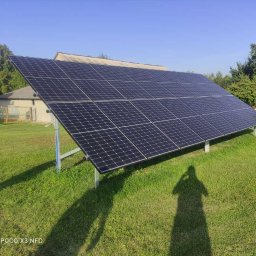 Renovatech sp. z o.o. - Instalacja fotowoltaiczna na trawniku, cień osoby fotografującej widoczny na pierwszym planie, budynek w tle, błękitne niebo.