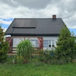 Dom jednorodzinny z panelami fotowoltaicznymi na dachu. Widoczne panele w czarnej ramie na tle szarej dachówki i błękitnego nieba z chmurami.