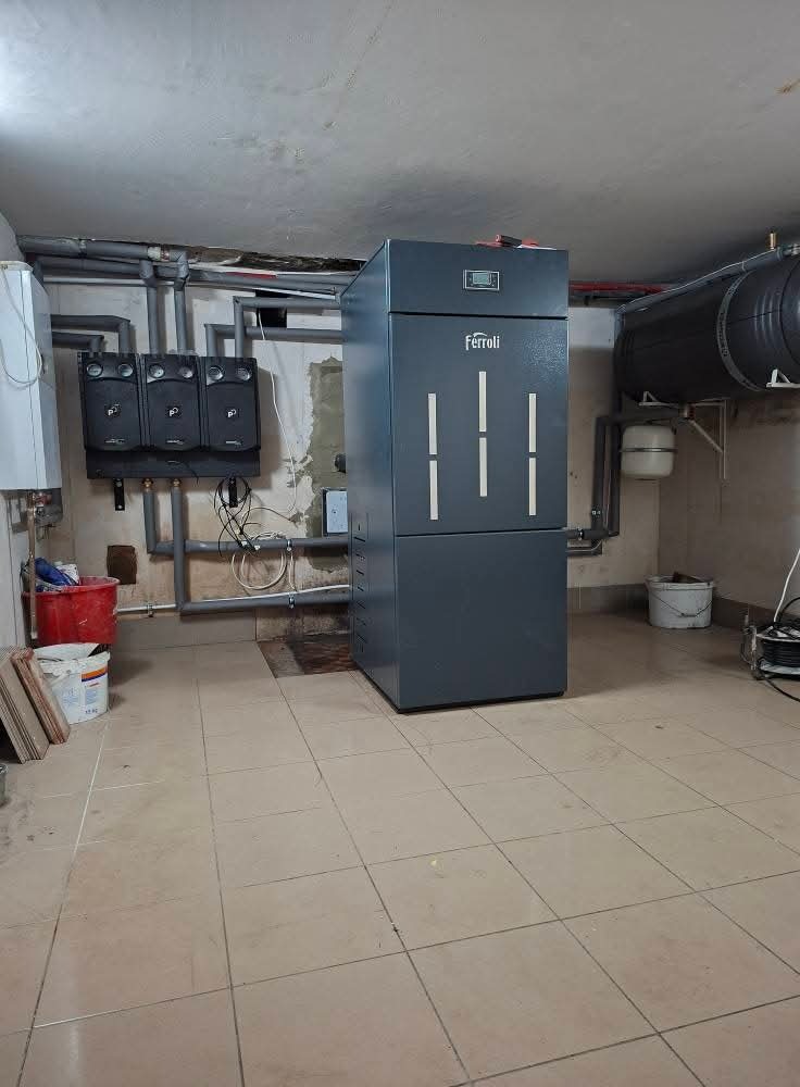 Szary piec Ferroli w kotłowni z widocznymi rurami, zbiornikiem wyrównawczym i panelami sterowania, na tle ceramicznej podłogi w beżowym odcieniu.