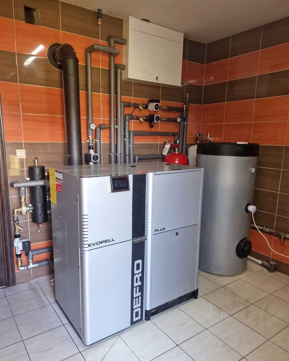 Kocioł Defro Evopell Plus z zasobnikiem w kotłowni wykończonej płytkami. Widoczne rury instalacji CO i CWU, armatura. Gotowa instalacja grzewcza.