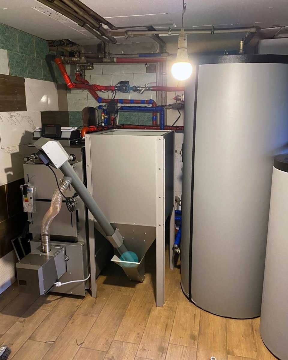 Nowoczesny piec na paliwo stałe z podajnikiem i zasobnikiem, zainstalowany w kotłowni z ceramiczną okładziną i drewnopodobną podłogą. Widoczne rury instalacji grzewczej.