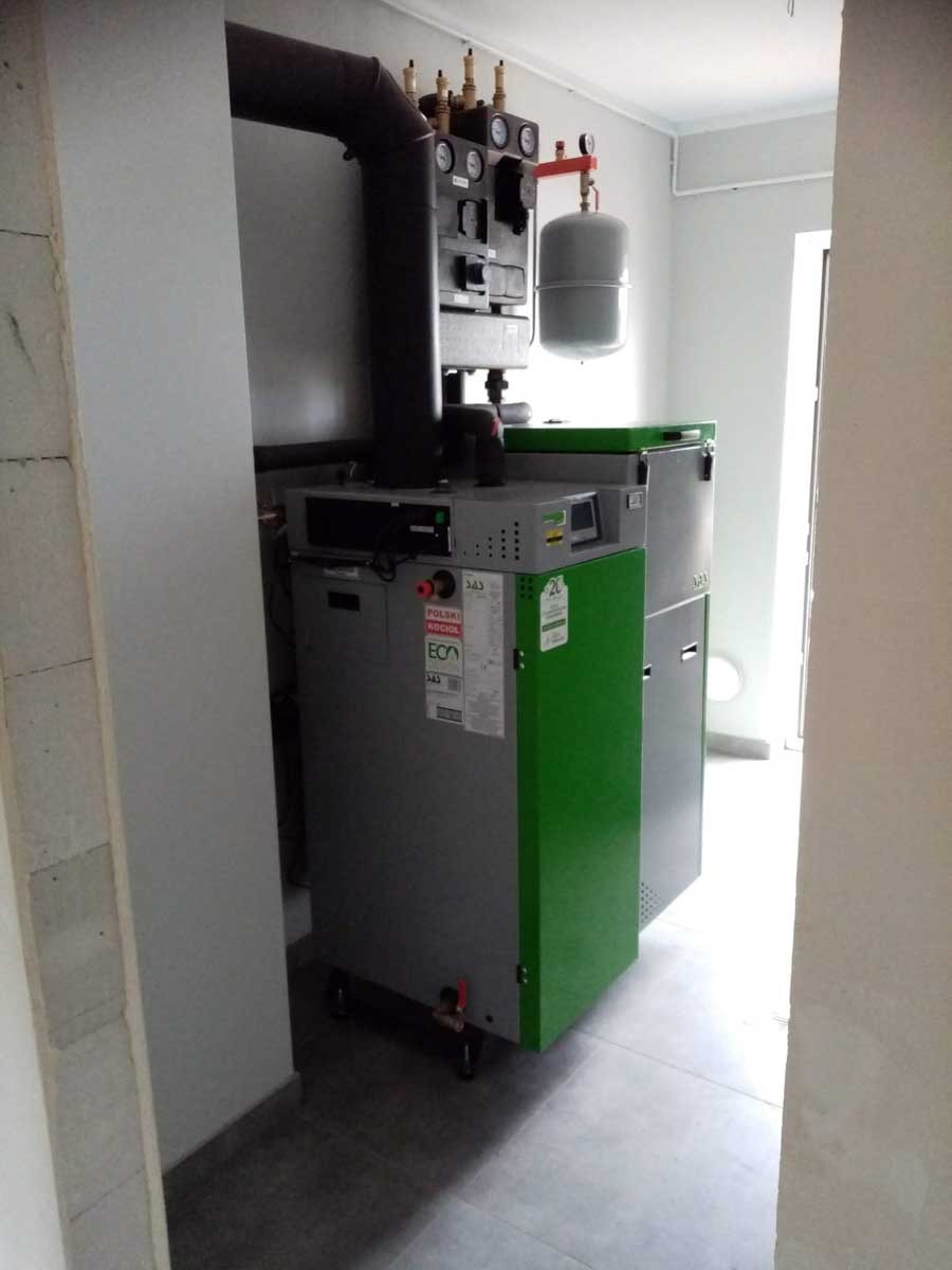 Szaro-zielony piec na paliwo stałe ECA w kotłowni z szarą instalacją. Widoczne elementy sterujące i rury. Ujęcie w pomieszczeniu z betonową ścianą.