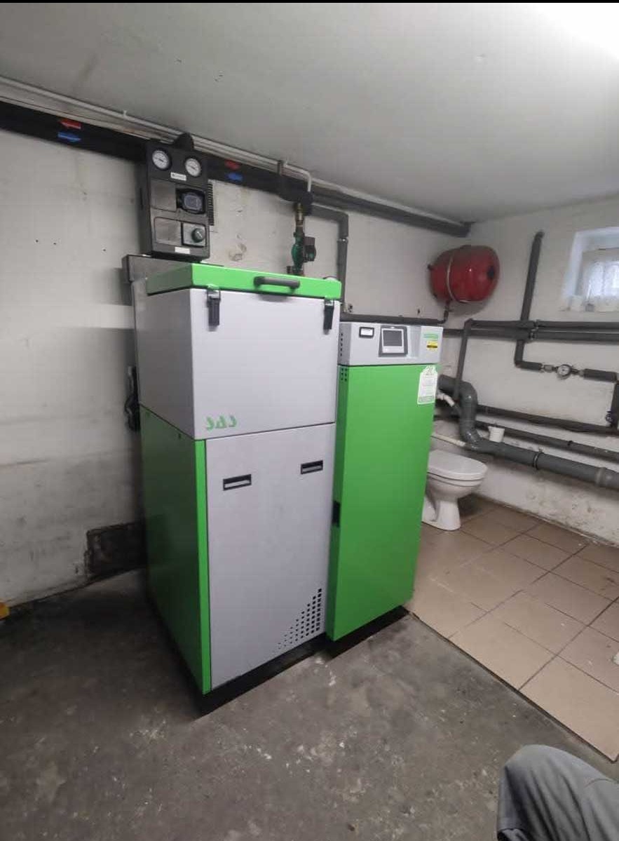 Szaro-zielony piec SAS w piwnicy z instalacją CO i bojlerem. Widoczne rury, manometry i fragment toalety. Ujęcie z dołu.