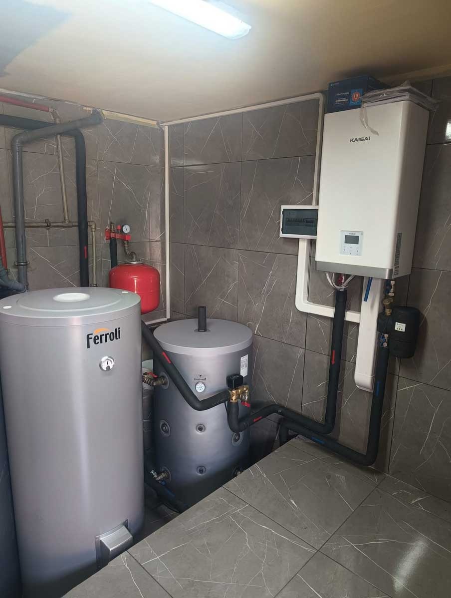 Kompaktowa kotłownia z zasobnikami Ferroli i Kaisai, rury w izolacji, szare płytki na ścianach i podłodze. Instalacja grzewcza w pomieszczeniu technicznym.