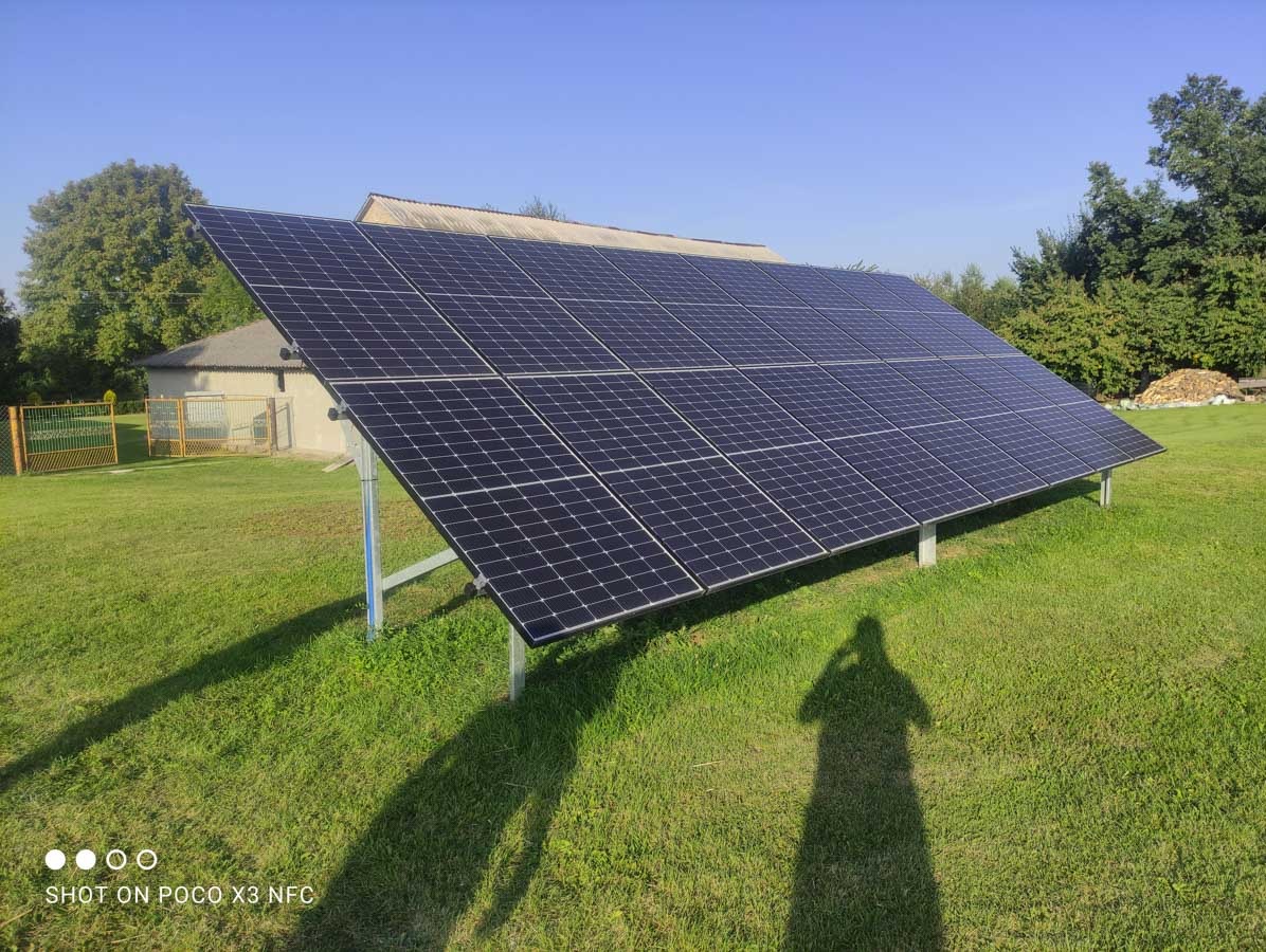 Instalacja fotowoltaiczna na trawniku, cień osoby fotografującej widoczny na pierwszym planie, budynek w tle, błękitne niebo.