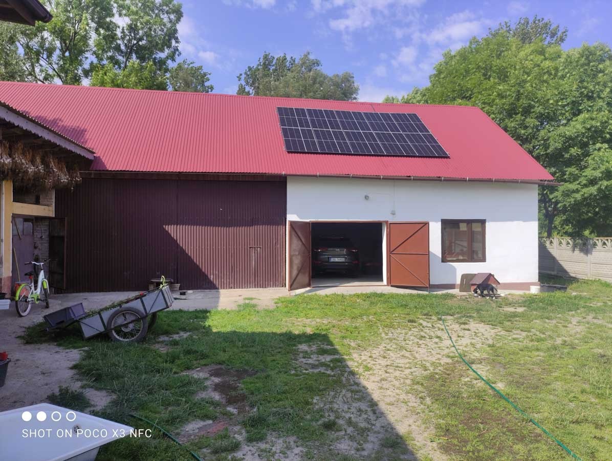 Biały budynek gospodarczy z czerwonym dachem i panelami słonecznymi. Otwarta brama garażowa, w tle zielone drzewa i błękitne niebo. Na pierwszym planie trawa.