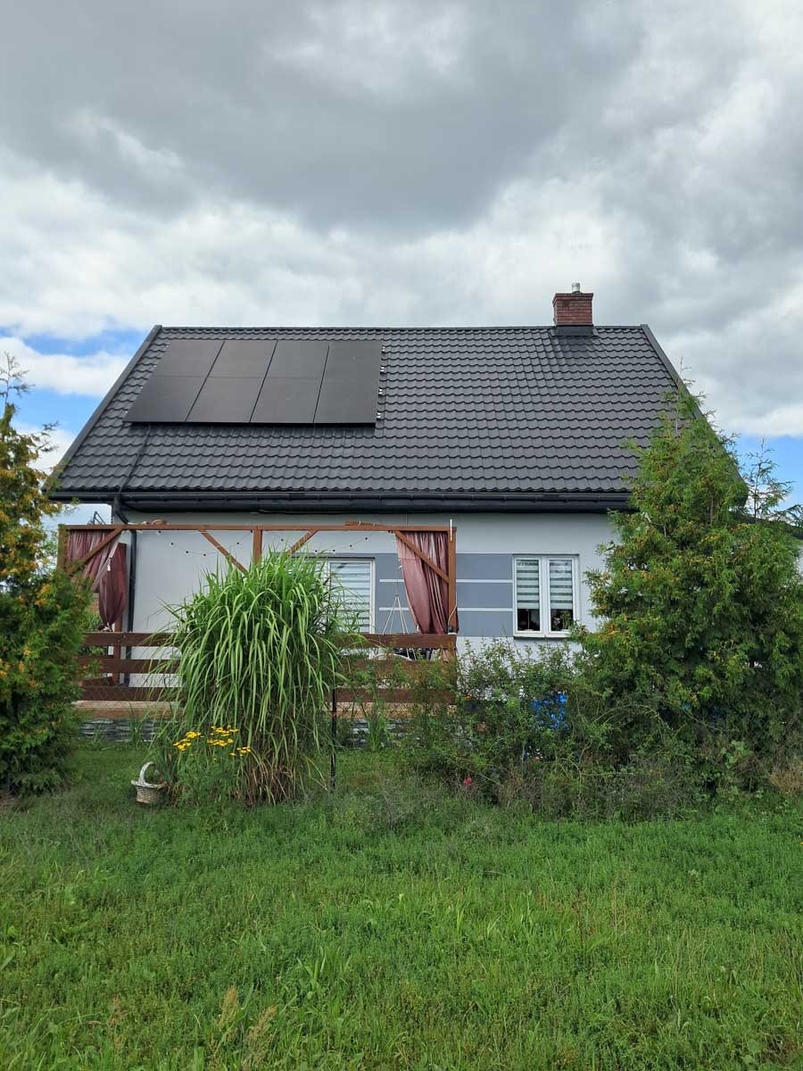Dom jednorodzinny z panelami fotowoltaicznymi na dachu. Widoczne panele w czarnej ramie na tle szarej dachówki i błękitnego nieba z chmurami.