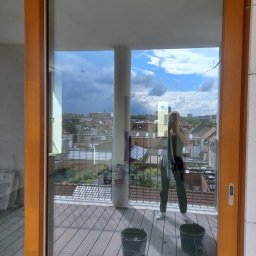 Katarzyna Klejna - Balkon z drewnianą podłogą, widok przez duże okno przesuwne. Dwie wiaderka i narzędzie do mycia podłogi sugerują świeżo zakończone sprzątanie.