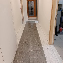 Katarzyna Klejna - Wypolerowana, szara posadzka w korytarzu biurowym w Toruniu. Widok na czyste, błyszczące płytki i drzwi w tle. Dbałość o detale i wysoki standard.