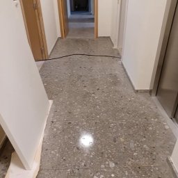 Katarzyna Klejna - Korytarz po sprzątaniu z terazzo, widoczne drzwi i kabel na podłodze. Świeżo umyta podłoga z połyskiem. Fragment drzwi oparty o ścianę zabezpieczony kartonem.