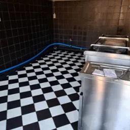 Wnętrze z czarno-białą szachownicą na podłodze i ciemnymi kaflami na ścianach. Widoczne metalowe urządzenia i niebieski wąż. Oświetlenie sufitowe.