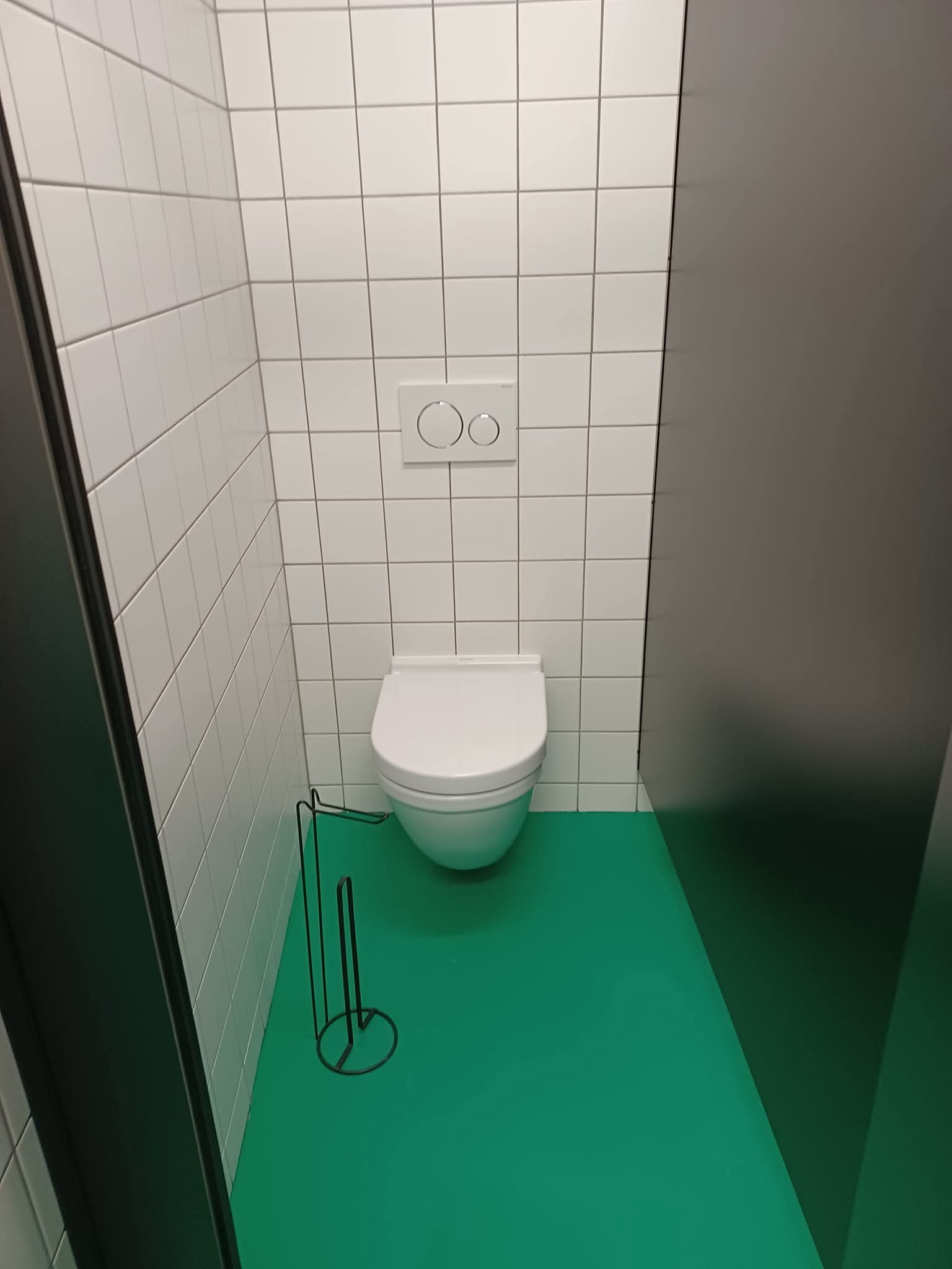 Minimalistyczna toaleta z białymi płytkami, zieloną podłogą i czarnymi akcentami. Widoczny uchwyt na papier toaletowy i panel spłuczki. Czyste, nowoczesne wnętrze.