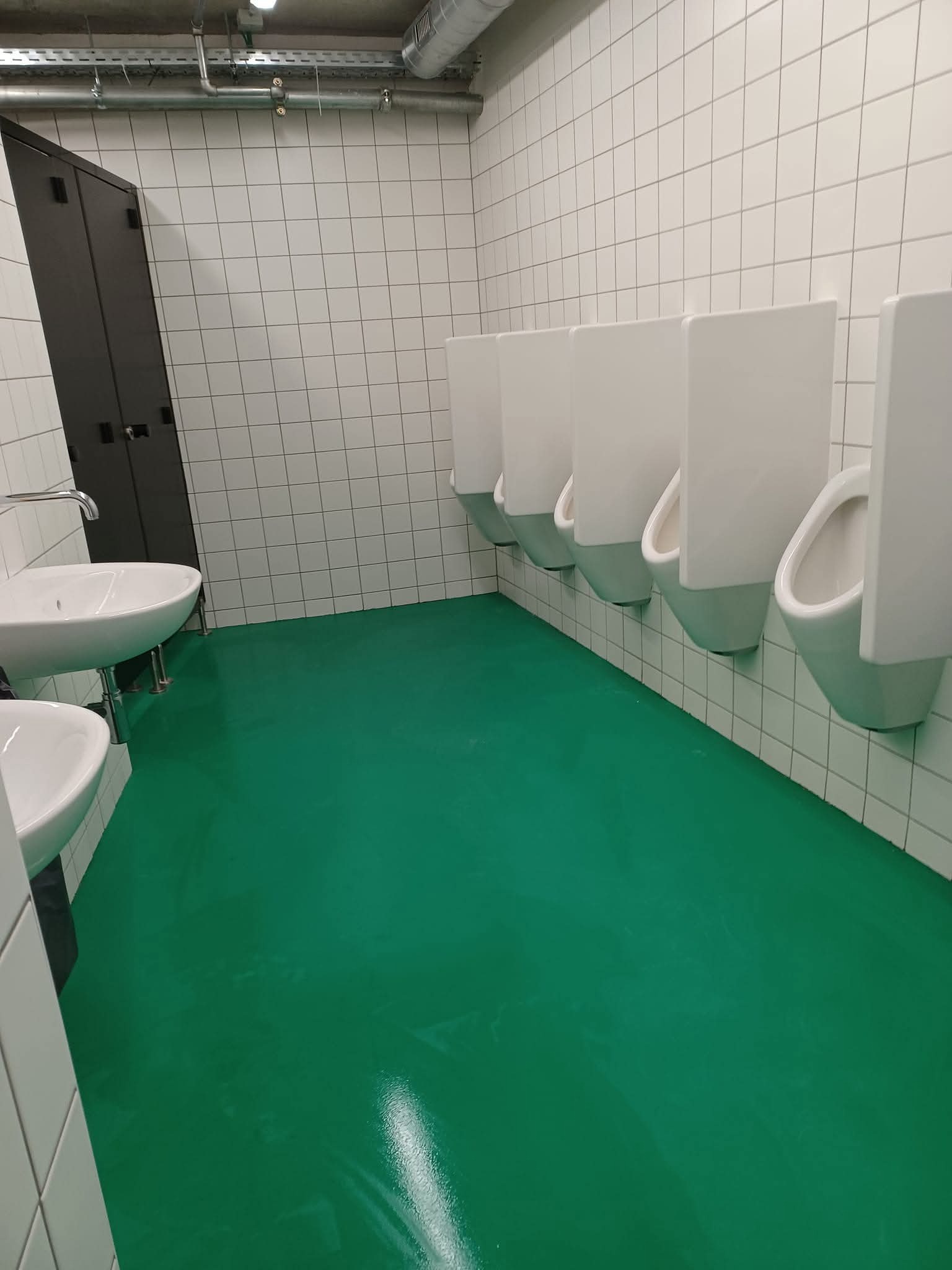 Wnętrze toalety publicznej po czyszczeniu: lśniąca, zielona podłoga, białe umywalki i pisuary, białe kafelki na ścianach, ciemna szafka z lewej strony.