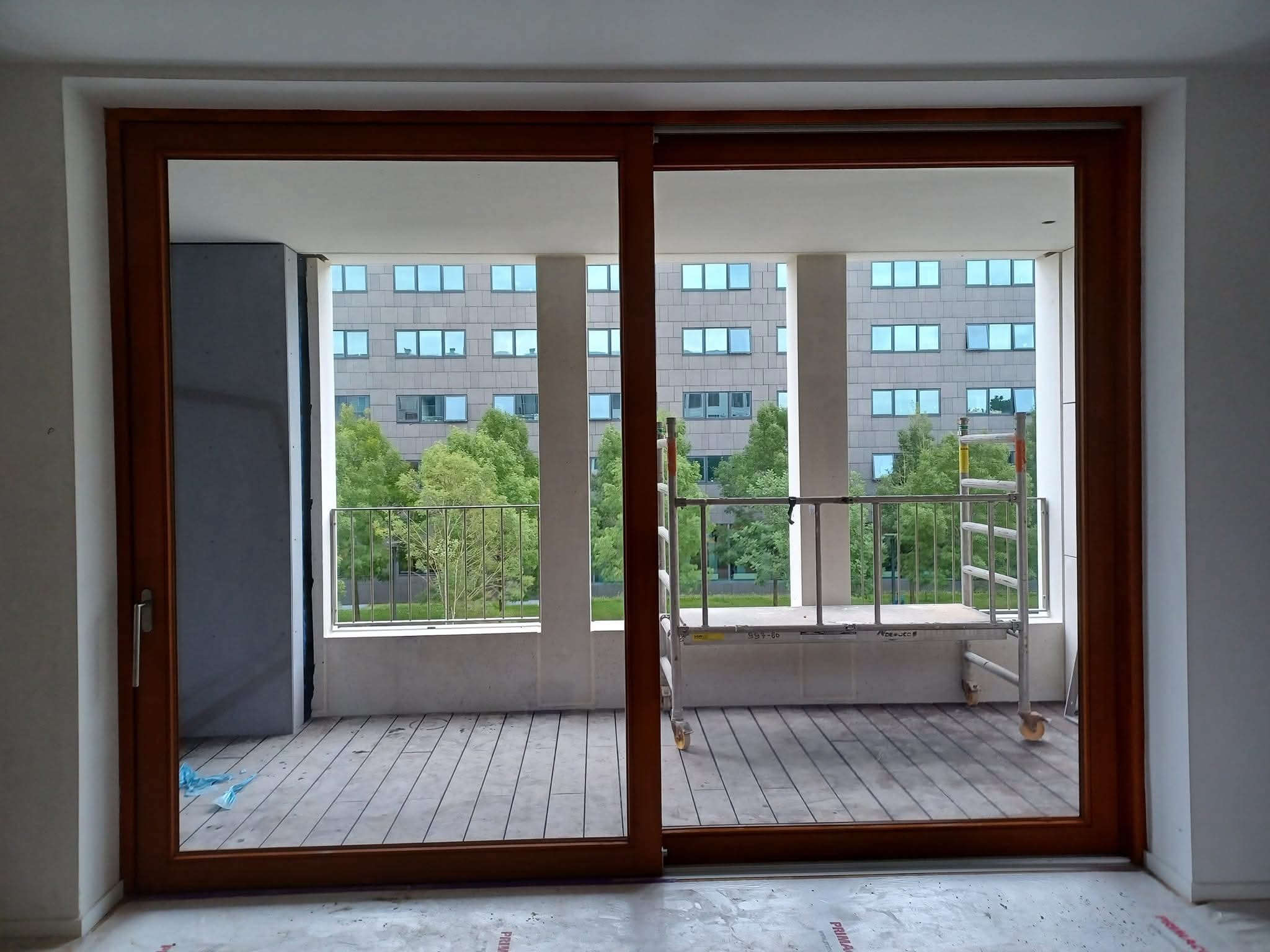 Duże, przesuwne okno balkonowe z drewnianą ramą, widok na balkon z drewnianą podłogą i rusztowaniem, w tle budynek z wieloma oknami.