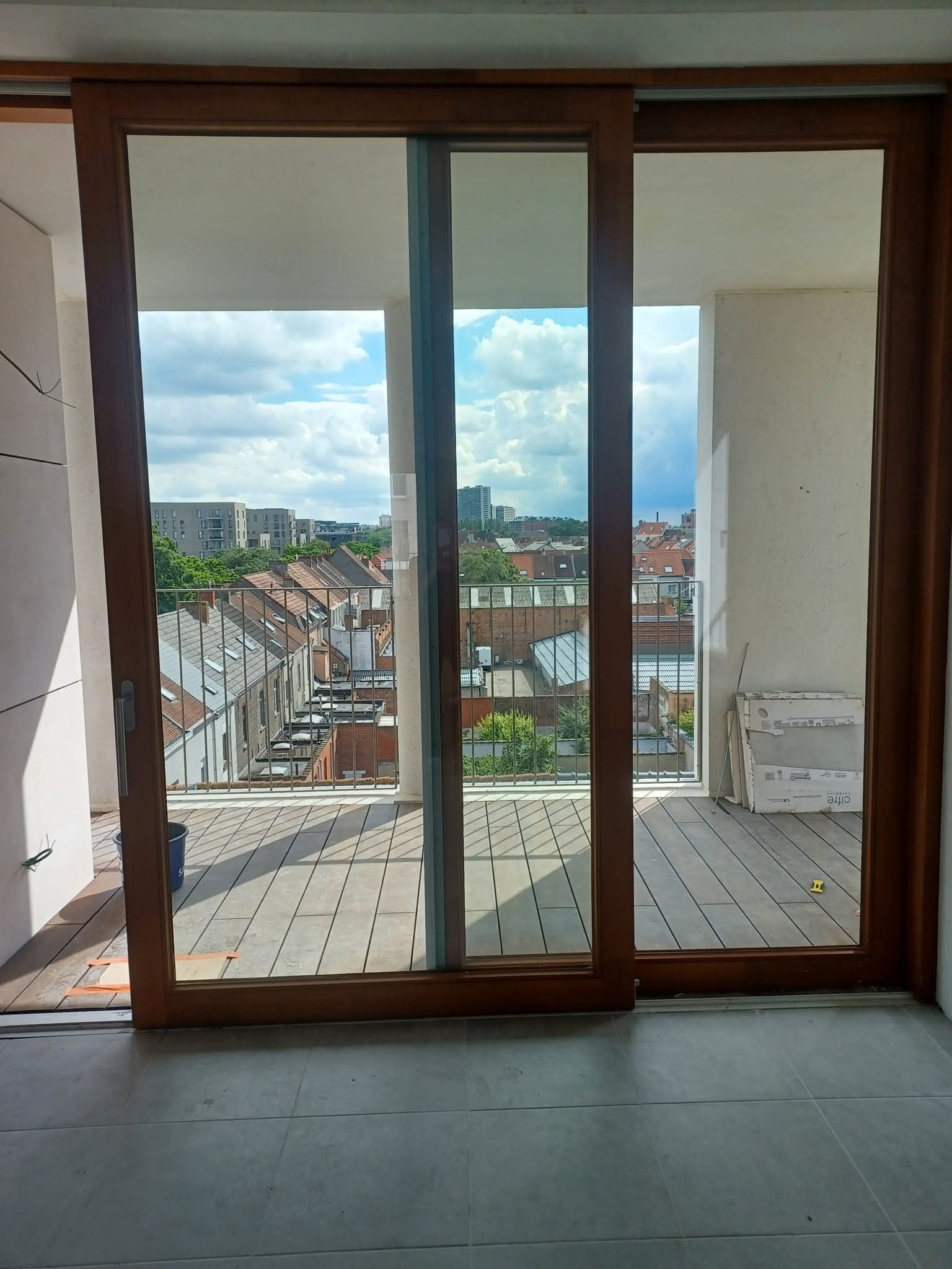 Widok z balkonu przez przesuwne okno balkonowe na zabudowę miejską w słoneczny dzień. Na balkonie leżą płyty i stoi wiadro. Podłoga wyłożona szarymi płytkami.