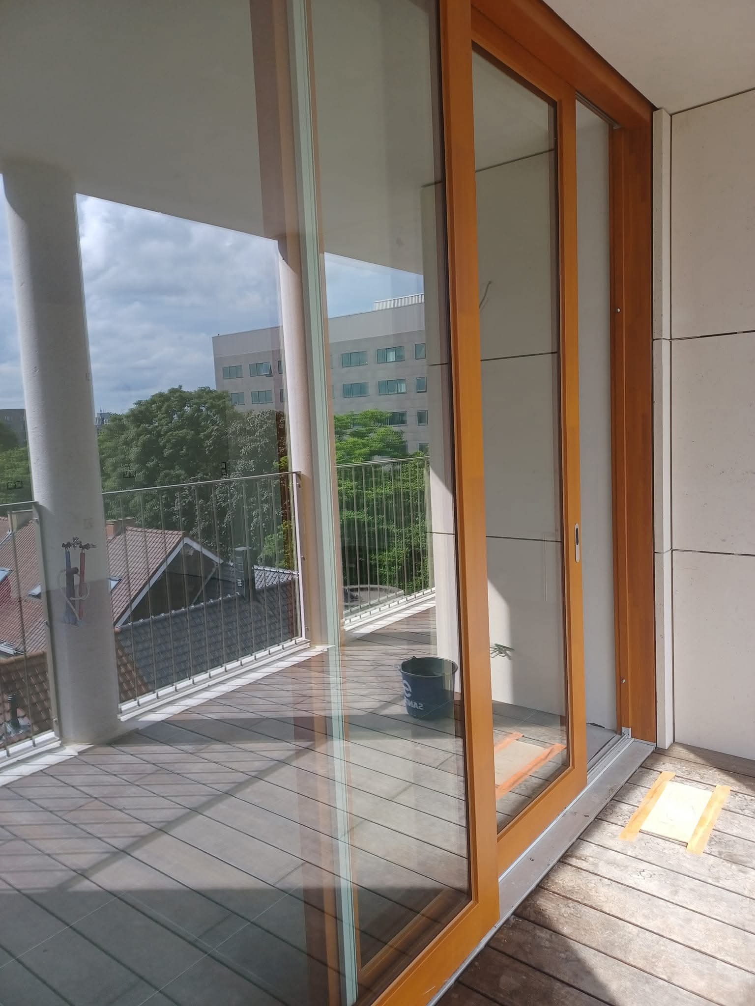 Balkon z drewnianą podłogą i przesuwnymi drzwiami. Widoczne wiadro, prawdopodobnie po myciu. Za barierką widok na budynki i zieleń.