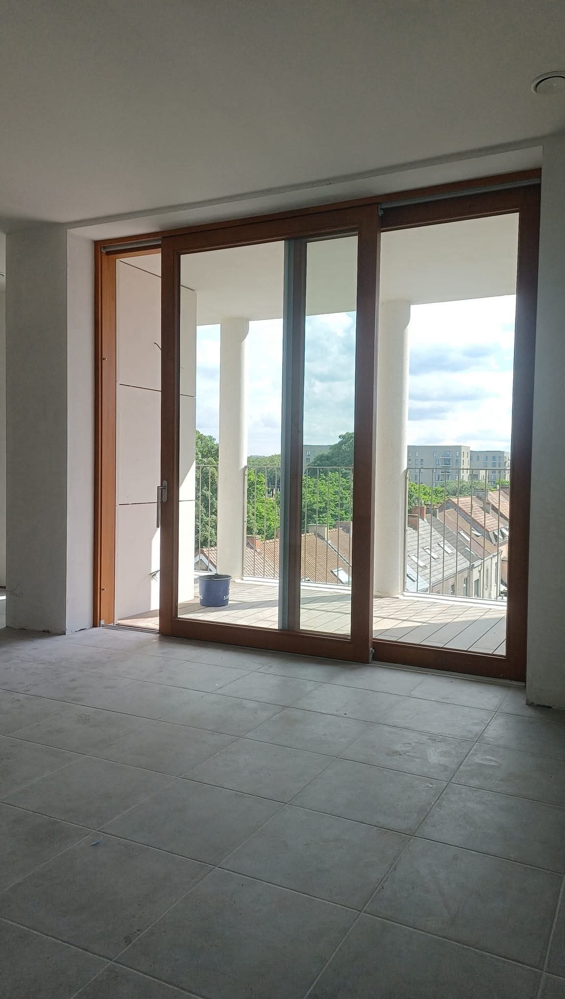 Wnętrze w stanie deweloperskim z dużym oknem balkonowym, widok na zieleń za balkonem. Na balkonie stoi wiadro. Szare płytki na podłodze, surowe ściany.