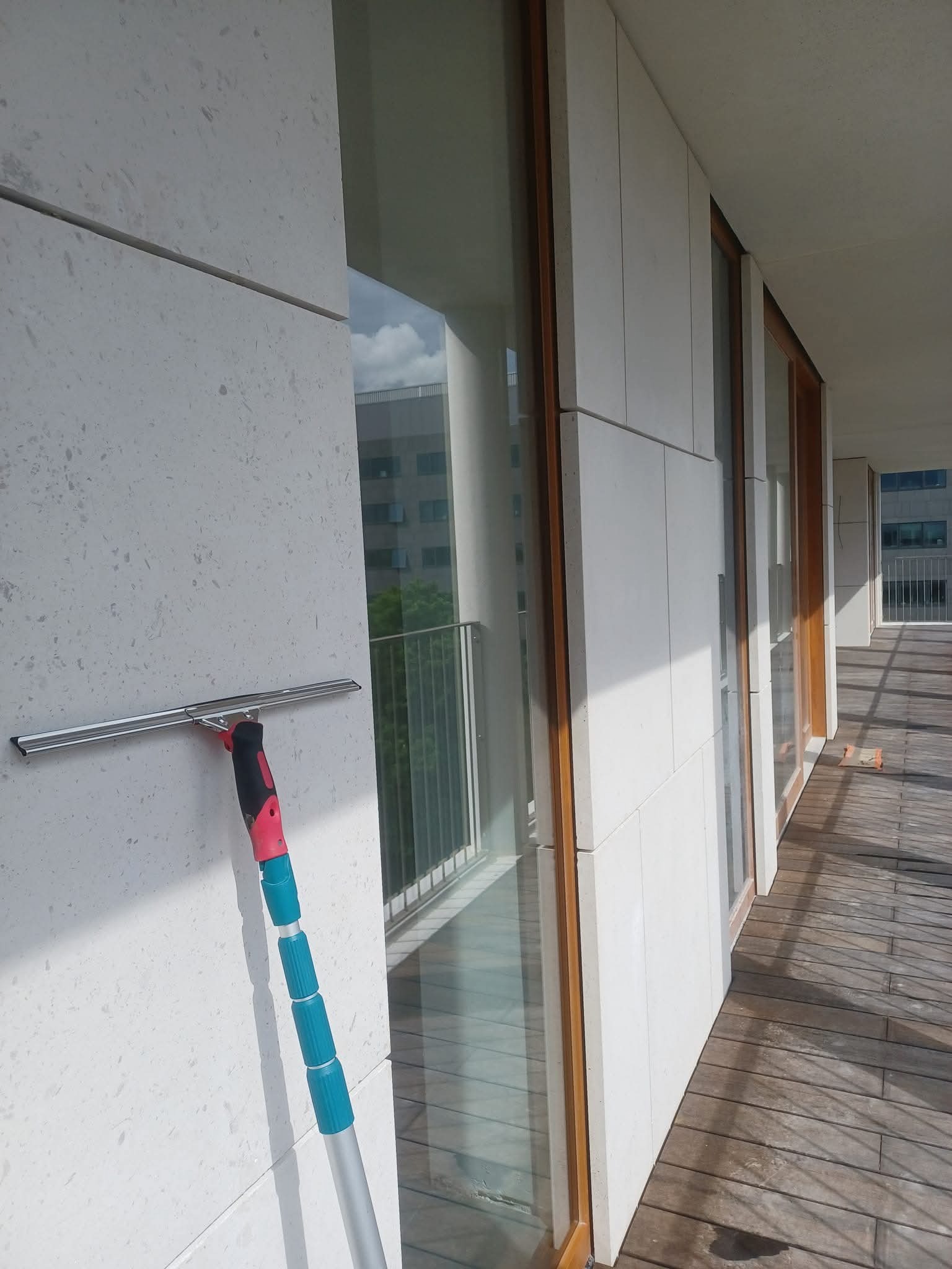 Srebrna myjka do okien z teleskopowym uchwytem oparta o elewację budynku. Widok na oszklony balkon z drewnianą podłogą w słoneczny dzień.