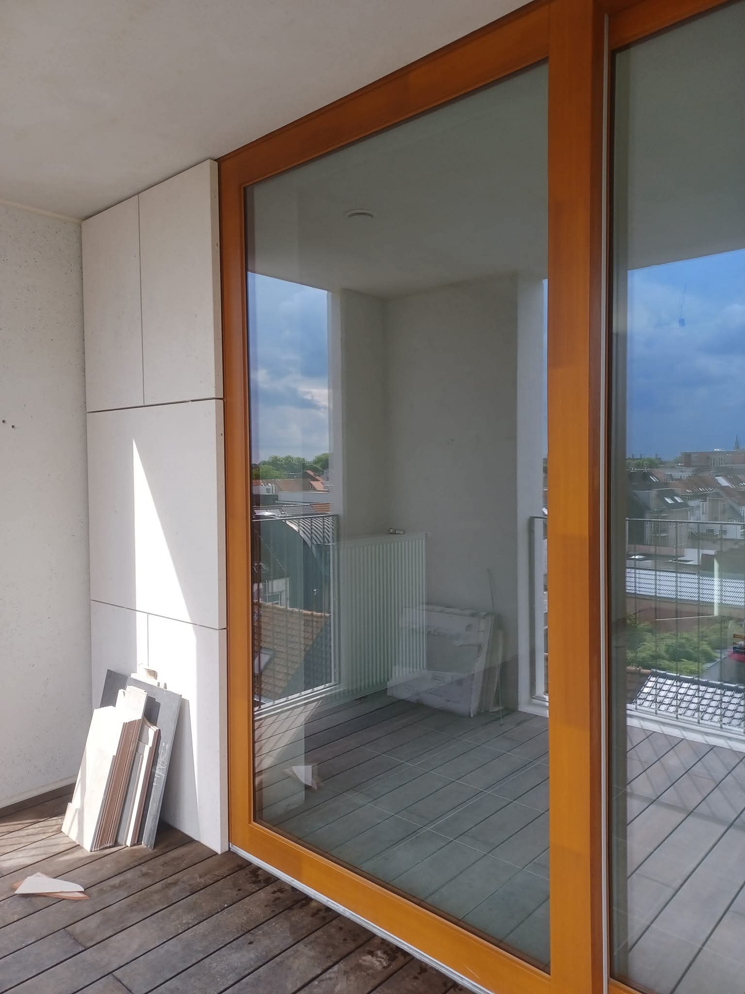 Balkon z drewnianą podłogą i dużym oknem przesuwnym. Przy ścianie leżą płytki, sugerujące prace wykończeniowe. Widok na budynki w tle.