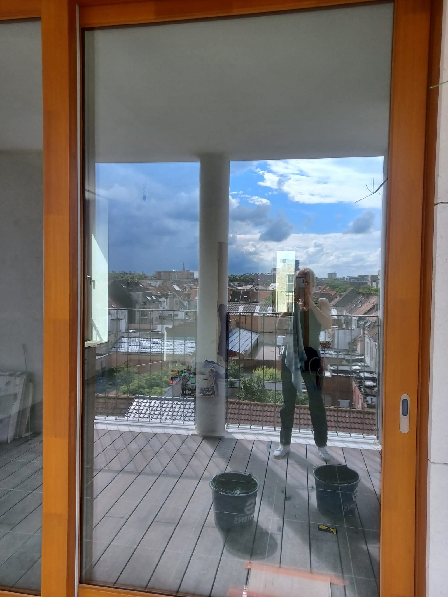 Balkon z drewnianą podłogą, widok przez duże okno przesuwne. Dwie wiaderka i narzędzie do mycia podłogi sugerują świeżo zakończone sprzątanie.