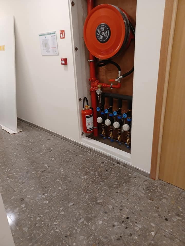 Wnęka z instalacją przeciwpożarową: zwinięty wąż, gaśnica i rury z zaworami. Szara posadzka z lastryko. Białe ściany i brązowe drzwi w tle.