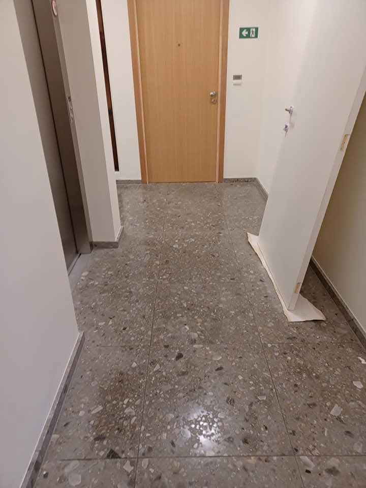 Korytarz z terazzo, widoczny fragment windy, drzwi wejściowe i białe drzwi opierające się o ścianę. Podłoga po umyciu, bez smug. Neutralne oświetlenie.