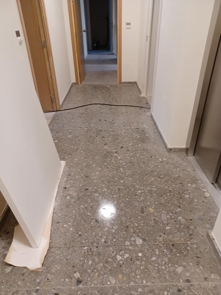 Korytarz po sprzątaniu z terazzo, widoczne drzwi i kabel na podłodze. Świeżo umyta podłoga z połyskiem. Fragment drzwi oparty o ścianę zabezpieczony kartonem.