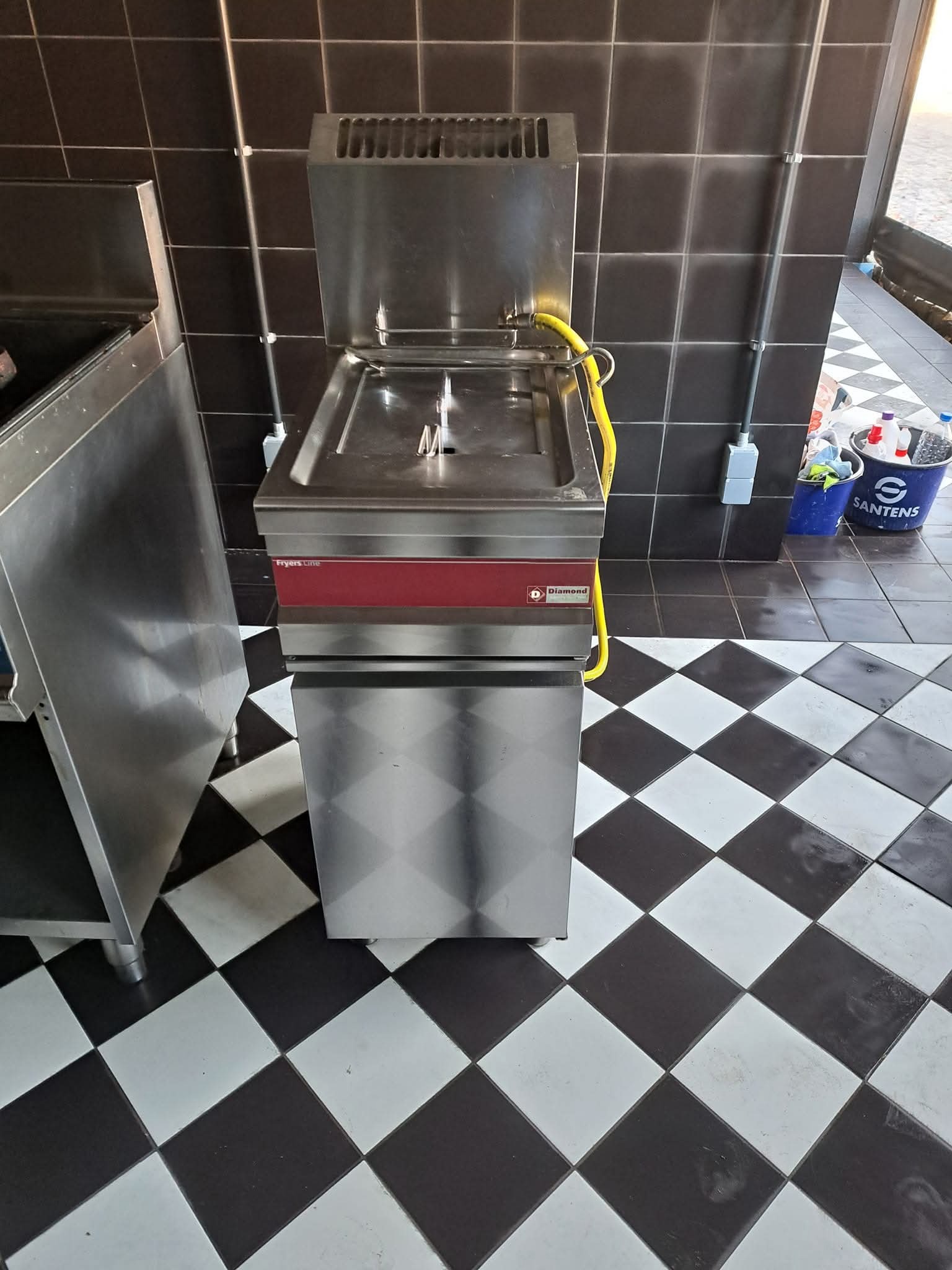 Profesjonalna frytownica Diamond z otwartą pokrywą, żółtym wężem i wiaderkiem Santens na czarno-białej podłodze w szachownicę i ciemnych kafelkach.