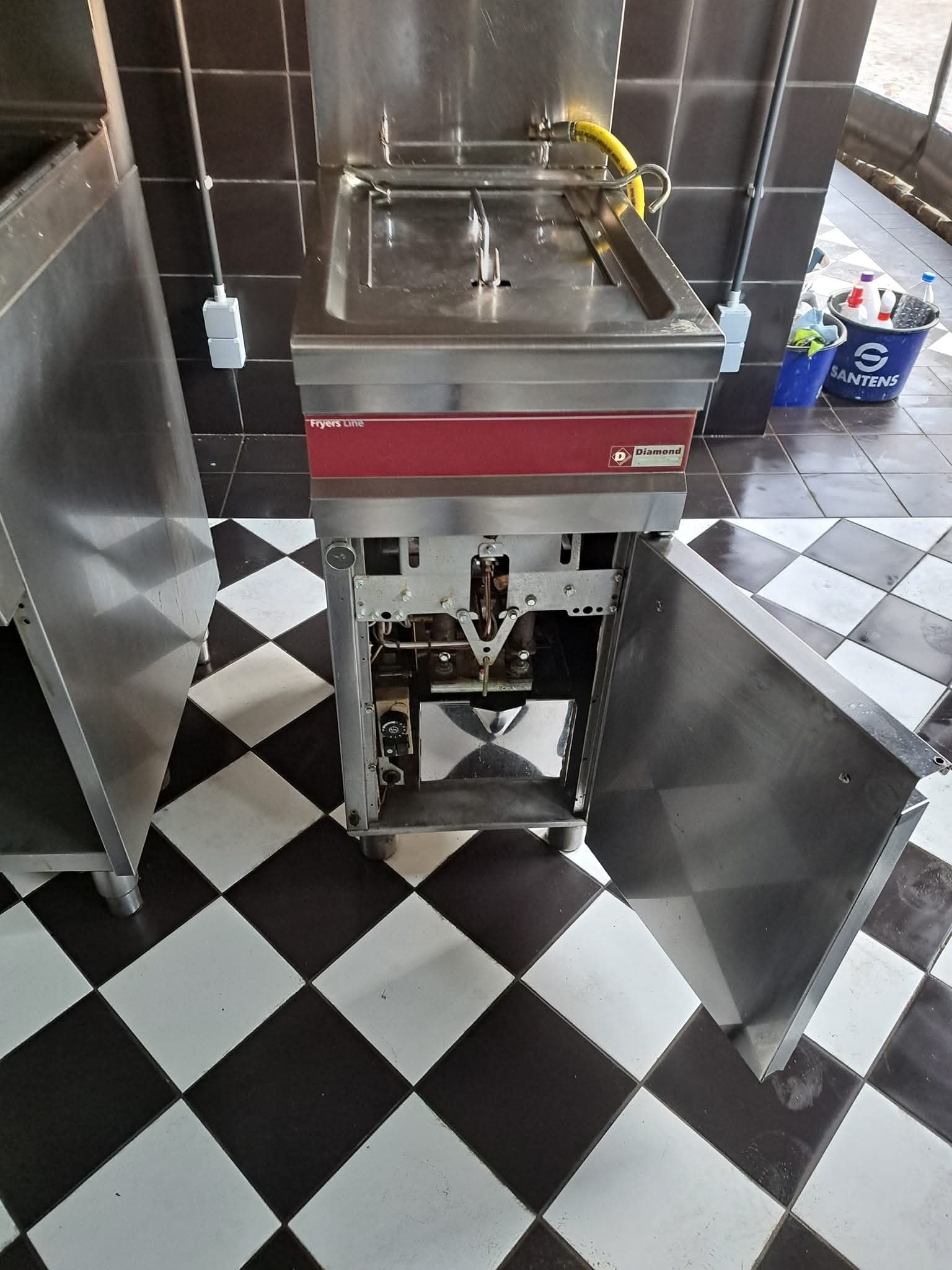 Otwarta, przemysłowa frytownica Diamond na czarno-białej podłodze w szachownicę, obok widoczny pojemnik z detergentami i akcesoriami do sprzątania.